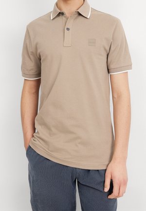 Man wearing beige short-sleeve polo shirt with white detail on collar and cuffs, paired with dark gray pants, standing with one hand in pocket.