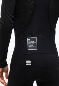 Ποδηλάτης που φοράει μαύρο μακρυμάνικο μπλουζάκι και σορτς bib με αντανακλαστικό λογότυπο και πίσω τσέπη που έχει λευκή ετικέτα με κείμενο ποδηλασίας.