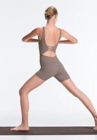Femme en combi-short marron à carreaux sans manches effectuant une position jambes écartées sur un tapis de yoga, dos face au spectateur, bras pliés aux coudes.