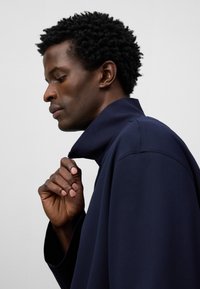 Marinblå tröja med hög krage, avslappnad passform och texturerad tyg. Modellen håller kragen något öppen, vilket visar skjortans design.