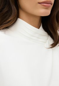 Top bianco con collo alto caratterizzato da pieghe stratificate vicino al collo, texture morbida e drappeggio delicato. Capelli parzialmente visibili.