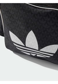 Črna tkaninska torba z velikim belim Adidas trefoil logotipom in vodoravnimi črtami, z teksturirano ozadje.