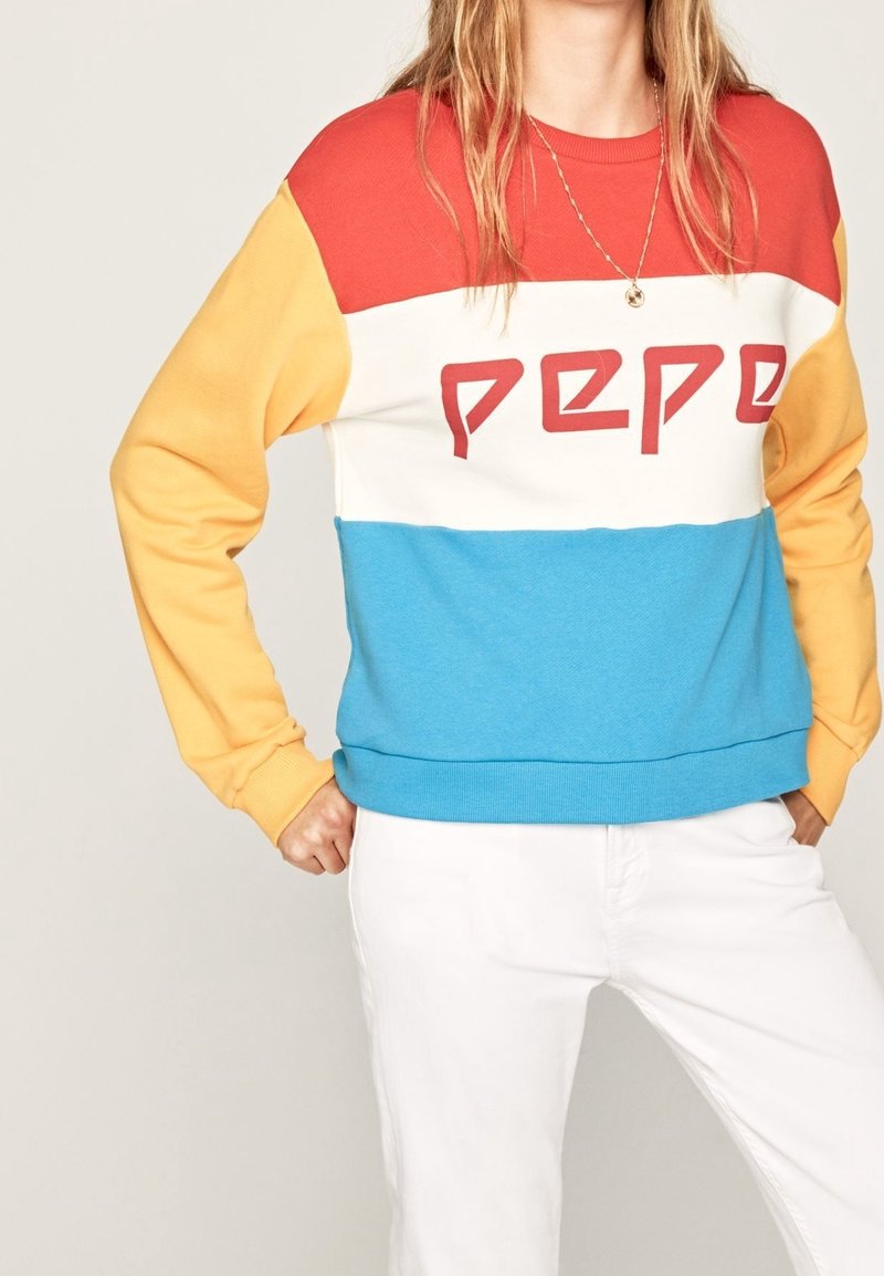Femme portant un sweat-shirt à panneaux de couleurs rouge, blanc, bleu et jaune avec un texte rouge et un pantalon blanc, debout les mains sur les hanches.