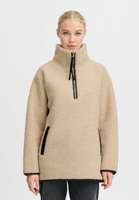Beige fleece pullover med høj krave, lynlås lukning og sorte accenter. Har sidelommer og en struktureret, blød stoffinish.