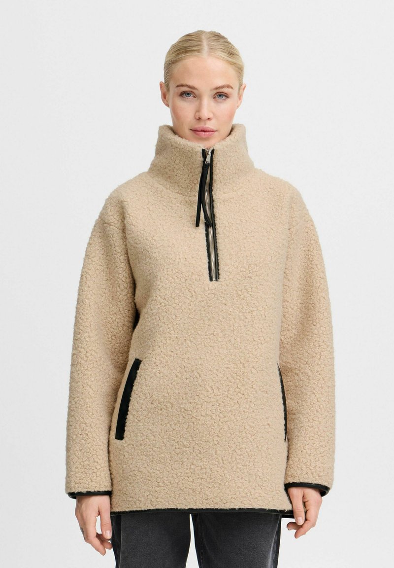 Beige fleece pullover med høj krave, lynlås lukning og sorte accenter. Har sidelommer og en struktureret, blød stoffinish.
