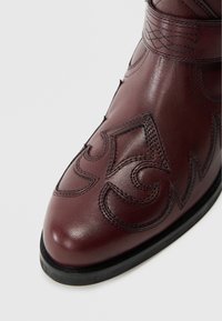 Vīna krāsas ādas zābaks ar sarežģītu melnu šuvi, smailu purngalu, teksturētām detaļām un zemu ādas zolīti.