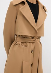 Trench-coat beige avec larges revers, boutons croisés, ceinture nouée et poche pliée, porté sur un top noir.