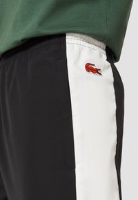 Svarta shorts med en vit sidpanel, prydda med en broderad röd krokodil-logotyp. Tillverkade av lättviktsmaterial, designade för komfort.