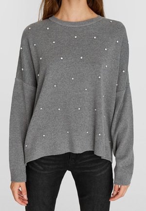 Vrouw die een losse grijze trui draagt met kleine witte kraaltjes verspreid over de voorkant, gecombineerd met een zwarte broek.