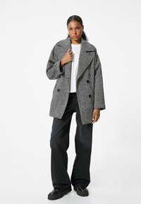 Szary płaszcz w wzór houndstooth, dwurzędowy, z szerokim kołnierzem i dwoma dużymi guzikami, połączony z czarnymi spodniami o szerokich nogawkach oraz masywnymi czarnymi botkami.