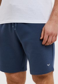 Shorts bleu marine en tissu doux, dotés d'une fermeture à cordon de serrage et d'un accent logo blanc. Le design comprend des ourlets cousus et une coupe décontractée.
