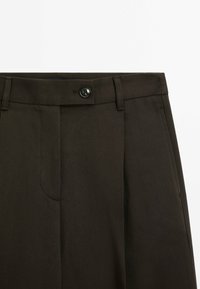 Gros plan sur un pantalon marron foncé avec une ceinture boutonnée, des passants de ceinture et des poches latérales sur fond blanc.