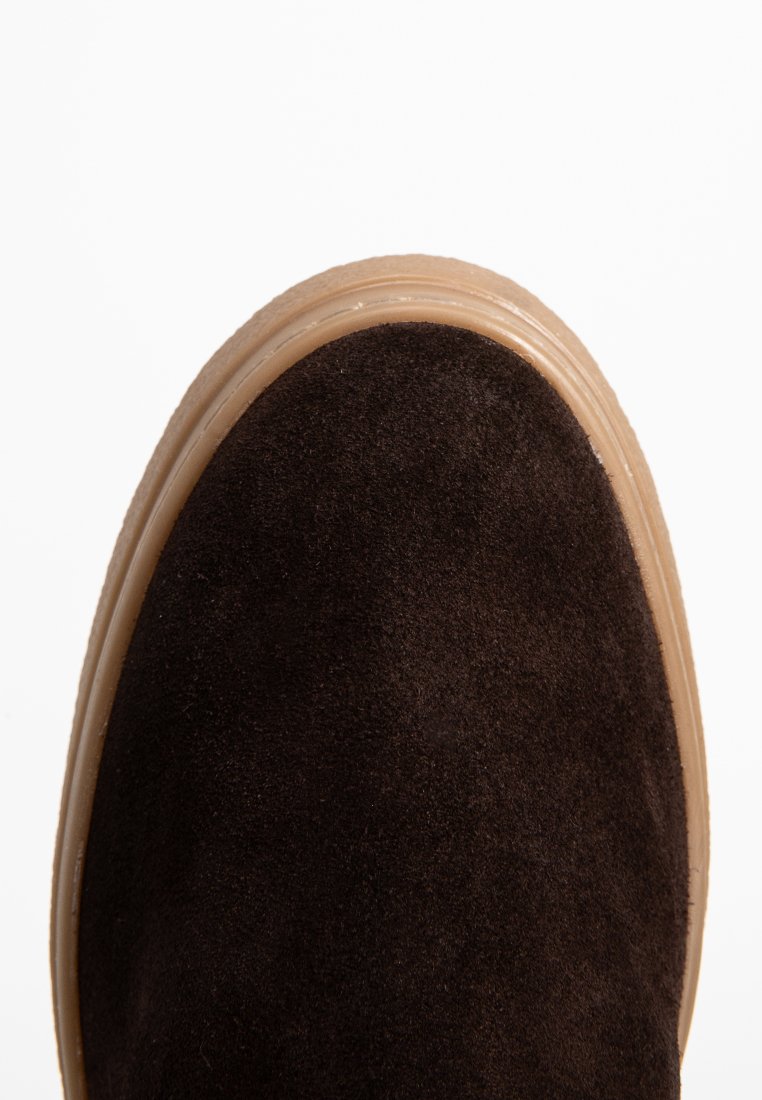 Dunkelbrauner Wildlederschuh mit beige Gummisohle auf weißem Hintergrund, der Textur und Nähdetails zeigt.