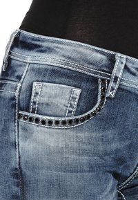 Jeans aus Denim mit einem verblassten blauen Finish, das ein einzigartiges Design der hinteren Tasche mit schwarzen Nietenakzenten und kontrastierenden Nähten aufweist.