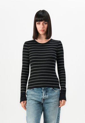Kvinne står mot en hvit bakgrunn iført en svart langermet skjorte med hvite horisontale striper og lyseblå jean.