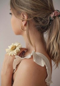 Orecchino ad anello attorcigliato in oro, elastico per capelli floreale, top crema con ruches. Fiore bianco appoggiato sulla pelle con un dettaglio texturizzato e centro verde.