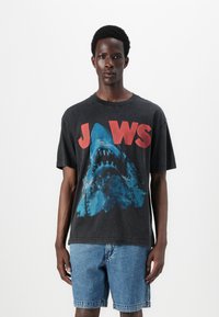 Svart grafisk T-skjorte med design av en blå hai og fet rød "JAWS"-tekst. Avslappet passform med korte ermer og rund hals. Brukt med denimshorts.
