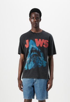 Sort grafisk t-shirt med et blåt hajdesign og fed rød "JAWS" tekst. Afslappet pasform med korte ærmer og rund halsudskæring. Bæres med denimshorts.