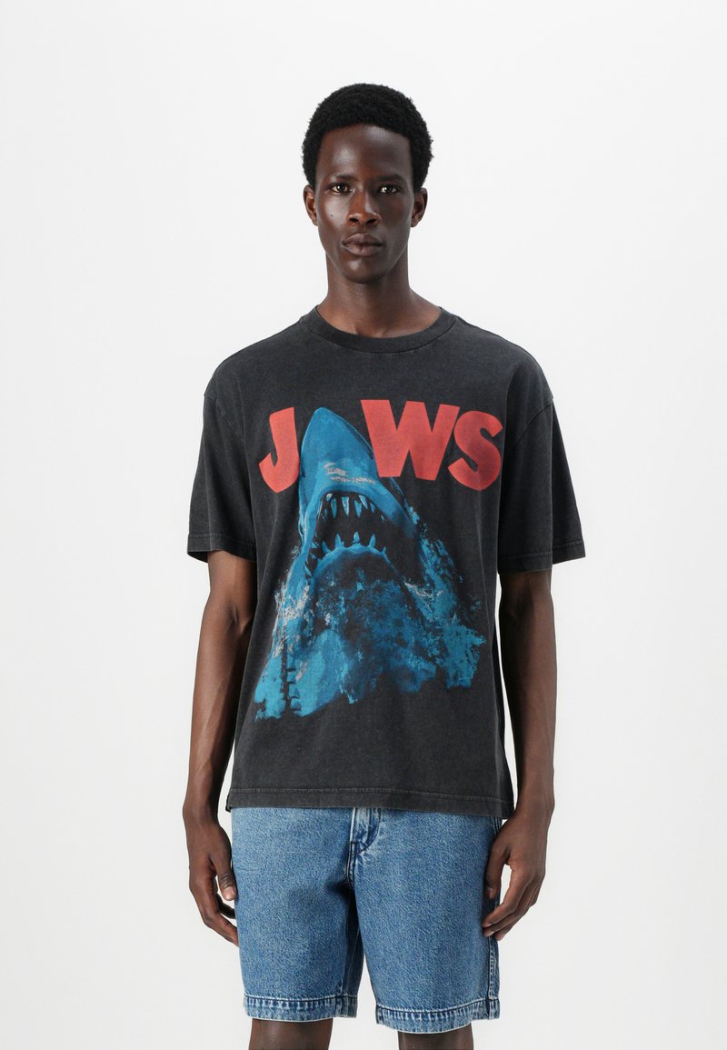 Svart grafisk T-skjorte med design av en blå hai og fet rød "JAWS"-tekst. Avslappet passform med korte ermer og rund hals. Brukt med denimshorts.