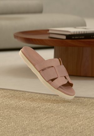 Ροζ σανδάλι τύπου slip-on με φαρδιές λουρίδες και λευκή σόλα τοποθετημένο σε μπεζ χαλί δίπλα σε στρογγυλό ξύλινο τραπέζι με βιβλία.
