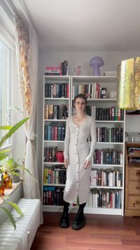 Vestido cinza claro com pregas e botões, de mangas longas, combinado com botas plataforma pretas. Prateleiras cheias de livros de várias cores ao fundo.