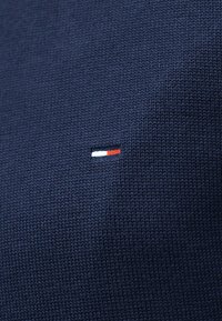 Tissu en maille bleu foncé avec un petit emblème rectangulaire brodé en blanc et rouge, centré sur la surface.