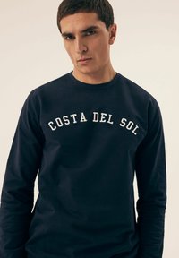 Granatowy długi rękaw sweter z okrągłym dekoltem i haftowanym napisem "COSTA DEL SOL" na białym tle na wysokości klatki piersiowej. Gładki materiał bawełniany.