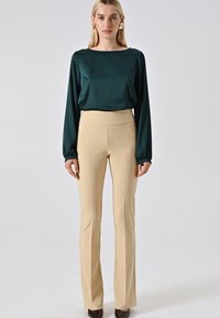 Donna che indossa una blusa a maniche lunghe verde scuro, pantaloni beige a vita alta, orecchini dichiarativi dorati e scarpe a punta marroni, su uno sfondo semplice.