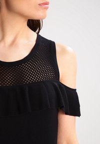 Top nero con scollatura in rete, tessuto a costine, design off-shoulder e volant lungo il bordo inferiore. La texture appare liscia.