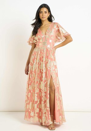 Woman wearing a coral maxi dress with gold floral patterns, flutter sleeves, V-neck, and a high slit, standing with one hand on hip.