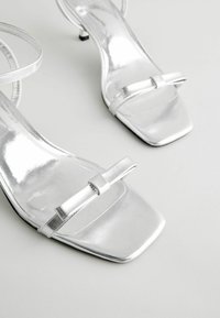 Silver high-heeled sandals with a glossy finish, square toe, and a decorative bow strap. Thin ankle straps and a small heel.