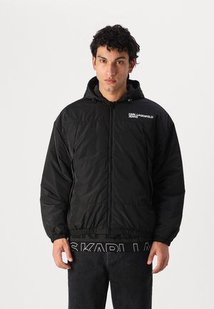 Schwarze Pufferjacke mit Kapuze, elastischen Bündchen und Saum. Verfügt über einen Frontreißverschluss und ein kleines weißes Logo auf der linken Brust.