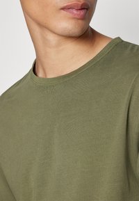 Oliwkowa koszulka z krótkim rękawem, wykonana z miękkiej bawełny. Posiada klasyczny dekolt i gładką fakturę z delikatnymi detalami szycia.