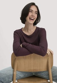 Junge Frau mit kurzen dunklen Haaren lächelt, während sie rückwärts auf einem Holzstuhl sitzt. Sie trägt ein lila langärmliges Oberteil und eine blaue Jeans.