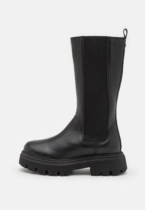 Botte en cuir noire mi-mollet avec semelle épaisse à crampons et panneau latéral élastique pour un enfilage facile, sur fond blanc.