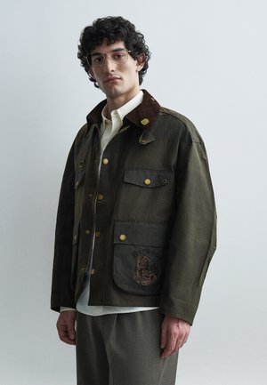 Muž s kučeravými vlasmi a okuliarmi má na sebe tmavozelenú bundu s hnedým golierom a vyšívaným vreckom, bielu košeľu a sivé nohavice.