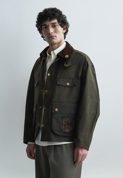Homme aux cheveux bouclés et portant des lunettes, vêtu d'une veste vert foncé avec un col marron et une poche brodée, d'une chemise blanche et d'un pantalon gris.