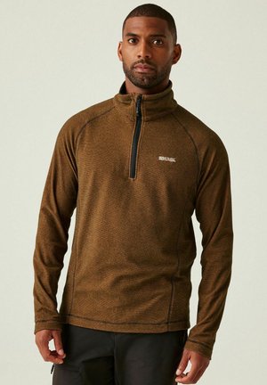 Man with short hair and beard wearing a brown quarter-zip pullover and black pants, standing against a plain light background.