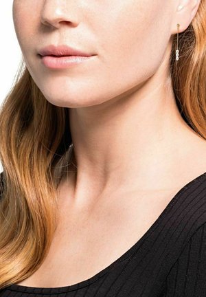 Goldene Ohrringe mit zarter Kette, die drei kleine weiße Perlen aufweisen, getragen von einer Person mit welligem roten Haar, in einem schwarzen gerippten Oberteil.