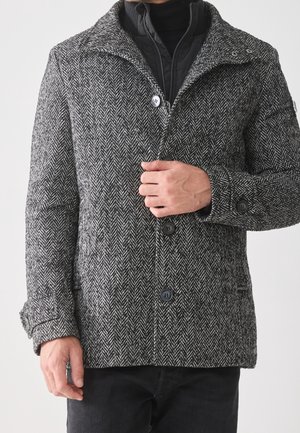 Homme portant un manteau en laine à motif chevron noir et blanc par-dessus une veste noire zippée et un pantalon noir.