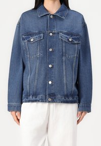 Jeansowa kurtka w średnim niebieskim kolorze o klasycznym kroju, z dwoma kieszeniami na piersi, srebrnymi guzikami i luźnym fasonem. Teksturowane wykończenie materiału.