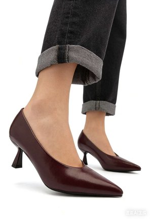 Pieds chaussés de chaussures à talons kitten pointues de couleur bordeaux foncé, associés à un jean noir retroussé, sur fond blanc.