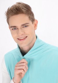 Hellesblaues Fleece-Weste mit Reißverschluss und hohem Kragen, glatter Textur und langen weißen Ärmeln. Das Model hält den Reißverschluss-Zipper.