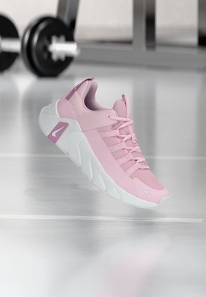 Lyserød og hvid atletik-sneaker med snørebånd, der svæver over et halmgulv med sløret vægtudstyr i baggrunden.