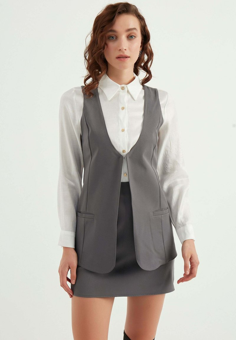 Grå väststyle topp över en vit skjorta med knappar, med en v-ringning, två framfickor och en figurnära silhuett.