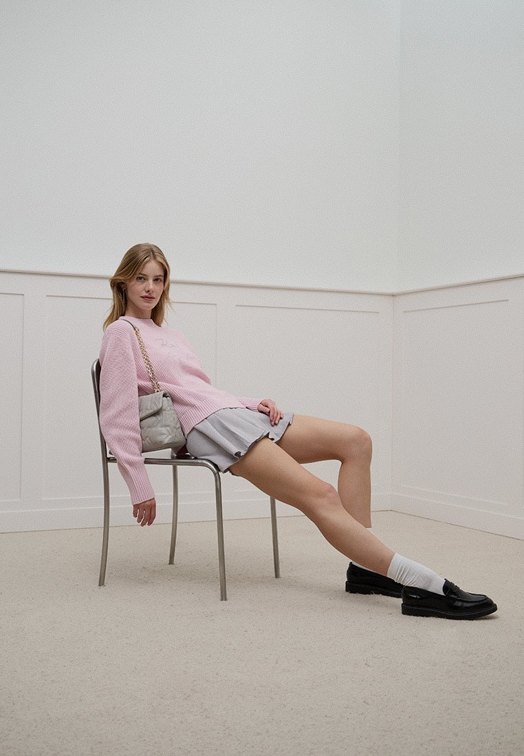 Pull en tricot rose, jupe plissée grise, chaussettes blanches et mocassins noirs. Modèle assis sur une chaise en métal dans un intérieur minimaliste.