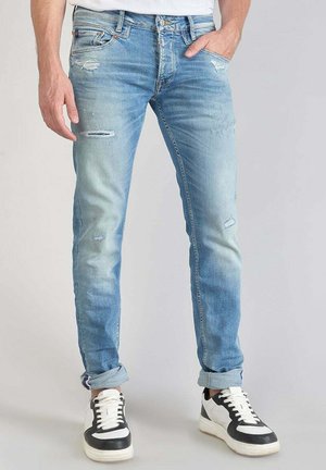 Jean en denim bleu clair avec une coupe slim, présentant des détails usés et une texture décolorée. Assortis à des baskets noires et blanches.