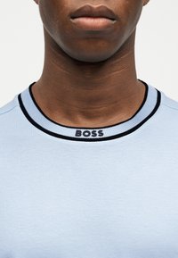 Κοντινό πλάνο του λαιμού και του κάτω μέρους του προσώπου ενός ατόμου που φοράει ένα ανοιχτό γαλάζιο πουκάμισο με ριμπ τσέπη με μαύρο περίγραμμα και τη λέξη "BOSS" σε μαύρο χρώμα.