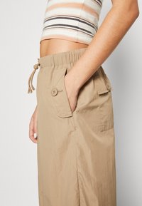 Beige, lichtgewicht cargobroek met een elastische tailleband, trekkoord, twee steekzakken aan de zijkant en knoopdetails. Gerimpelde textuur zorgt voor visuele aantrekkingskracht.