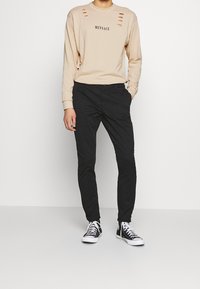 Beige mikina s dlouhým rukávem, potiskem a se zašlými výřezy, kombinovaná s černými slim-fit kalhotami a černo-bílými teniskami.